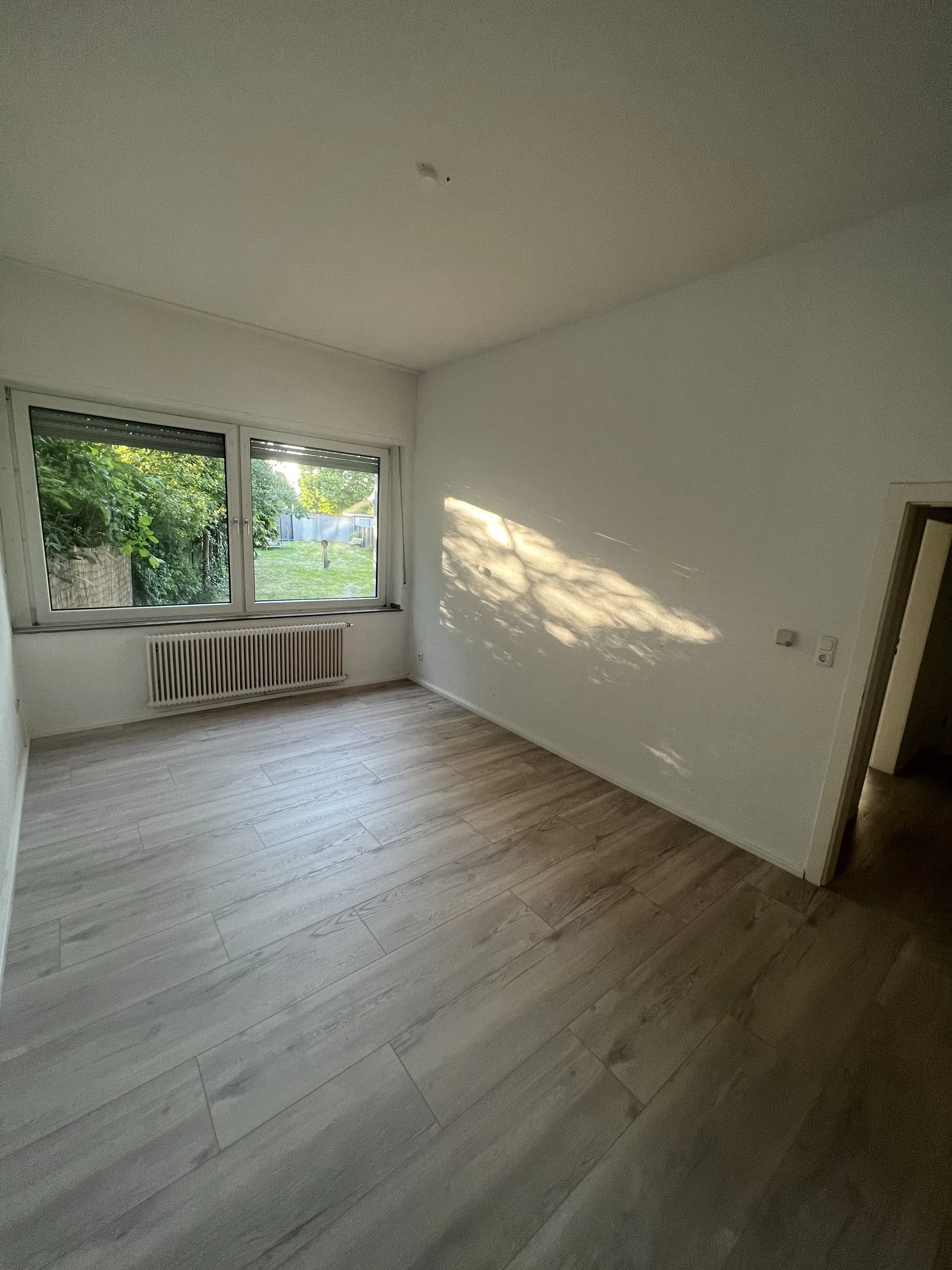 Heller Wohnraum mit Dielenoptik, zwei Fenstern zum Garten und Sonnenlicht auf der Wand